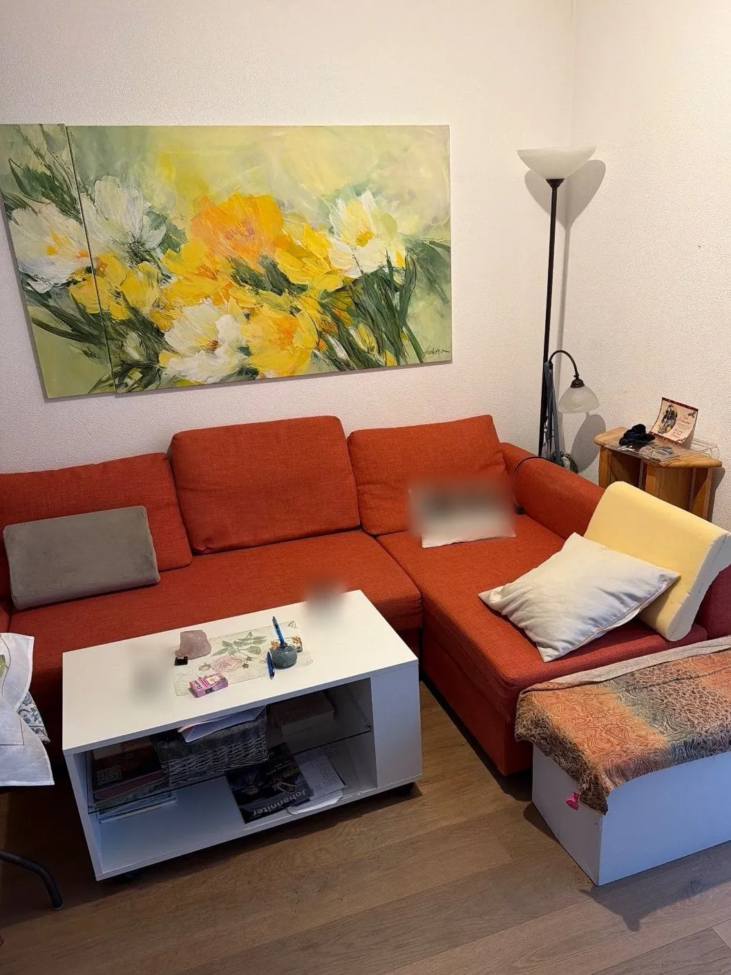 Wohnraumecke mit orangefarbenem Ecksofa, weißem Couchtisch, Stehlampe, kleinem Beistelltisch und großem Blumenbild an der Wand.
